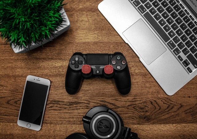 silver MacBook, black Sony ps4 dualshock4, silver iPhone 6 and round black keychain on brown wooden table